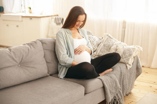 カジュアルな服を着た愛らしいかわいい若い妊婦は、彼女の腹に手を置き、彼女の胎児の息子と話し、幸せそうに微笑み、最初の妊娠の甘い瞬間を楽しんで、ソファに快適に座っています - comfortably ストックフォトと画像