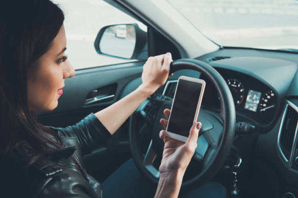 istock Young woman looking to her smartphone while driving car at front window background. Distracted female driver. Concept of dangerous driving. Woman driving and holding cellphone with blank screen 1139876286