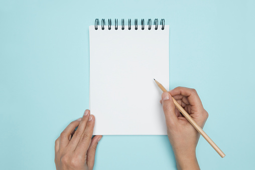 Hand is holding pencil and is about to write something into a spiral notepad on blue background.