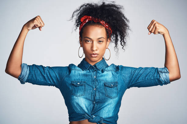 Look at these guns Portrait of a confident young woman wearing denim clothes and looking strong while flexing her biceps against a grey background girl power photos stock pictures, royalty-free photos & images