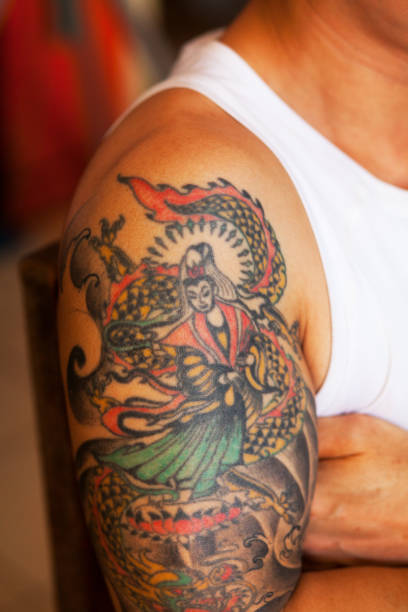 Tattoo on arm ad biceps of thai man - man is sitting in tanktop in front of house on sidewalk in street in Bangkok. Detailed shot of arm with tattoo. Tattoo is chinese style with a person in mandarin fashion style.