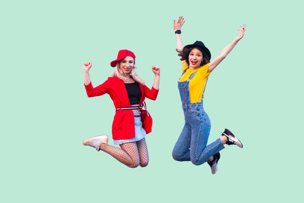Body size portrait of two happy screaming stylish hipster girls in fashionable clothes are jumping up in the air and looking at camera with raised arms. Body size portrait of two happy screaming stylish hipster girls in fashionable clothes are jumping up in the air and looking at camera with raised arms. Indoor studio shot,isolated on green background teenager couple child blond hair stock pictures, royalty-free photos & images