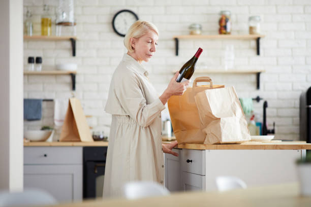 mature lady unpacking paper bags in kitchen - wine bottle wine residential structure alcohol imagens e fotografias de stock