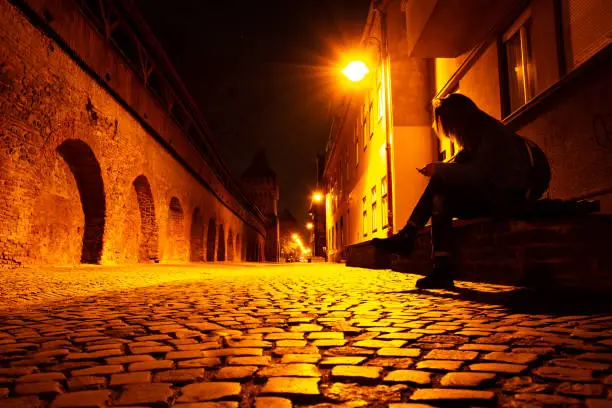 Young woman with a phone in hand, on a bench, late at night, on a medieval style street in Sibiu, Romania, with cobblestones lit warm yellow/red by street lamps, near a gothic wall with arches