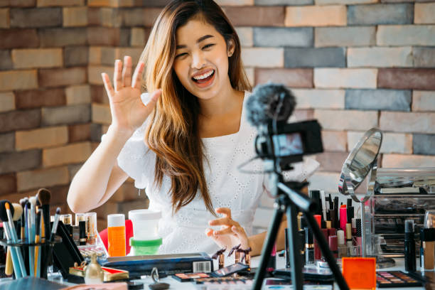 Smiling Asian woman waving hand on camera at table with cosmetics