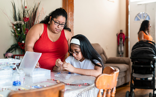 The body-positive, pretty Mexican Latino woman doing craft, fancy beadworks, together with her daughter, when her sister playing with the little boy in the room. Pennsylvania, USA.