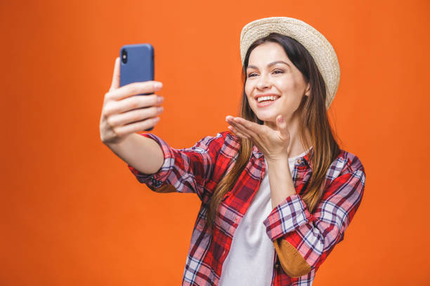 istock Young cheerful attractive brunette is smiling on the orange background. She is taking selfie on the camera of her phone, wearing casual summer outfit and a hat. 1139920678