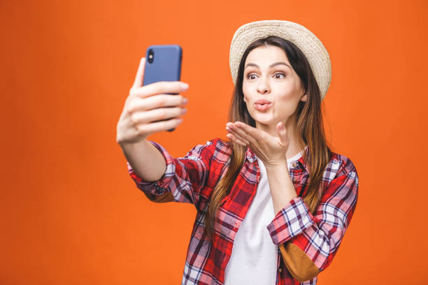 istock Young cheerful attractive brunette is smiling on the orange background. She is taking selfie on the camera of her phone, wearing casual summer outfit and a hat. 1139920861