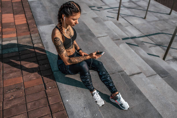 Athlete girl tattoos, sitting on steps summer city, hand smartphone, listening music with headphones. Resting after jogging workout. Concept of motivation, active life position healthy lifestyle. tatuajes chica atleta, sentado en los pasos de la ciudad de verano, teléfono inteligente de mano, escuchando música con auriculares. descansando después del entrenamiento de jogging. concepto de motivación, posición activa de la vida, vida sana.