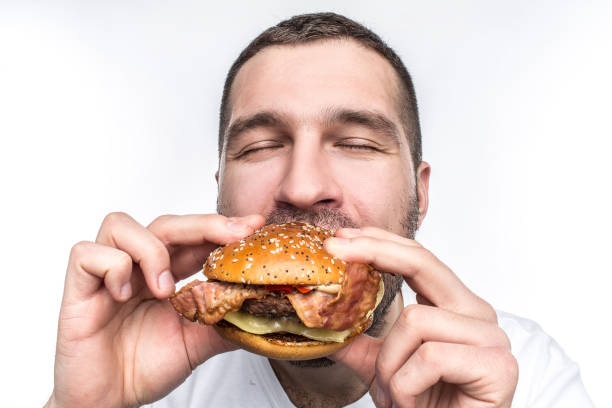 Weird and bizarre man is eating fat and juicy hamburger. It is not a healthy food but the guy likes it very much. His face is very emotional. Isolated on white background. stock photo