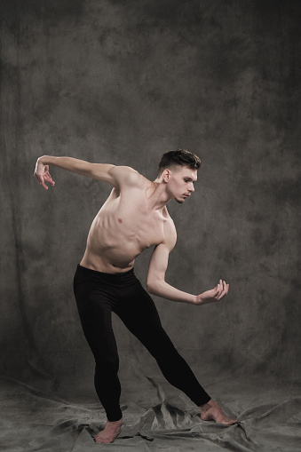 A young male ballet dancer with black leggings and a naked torso performs dance moves against a gray grunge background, with a light of lights and smoke. Conceptual art of contemporary.