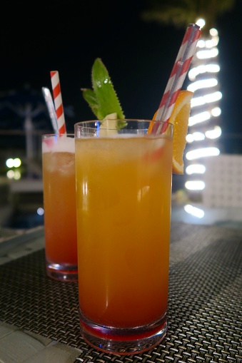 Photo showing non alcoholic orange ‘sex on the beach’ mocktails with grenadine in tall glasses at bar, outside al fresco dining with cocktail drinks at night, red and white straws, pineapple and orange garnish, garden lighting, spotlights and fairy lights wrapped around palm tree trunk.
