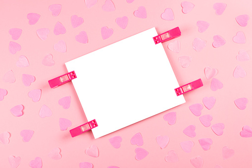 White blank sheet of paper fastened with clothes pins on cute and trendy pink background decorated with heart-shaped confetti. Copy space for text. Love message, Valentine's day and holiday concept.