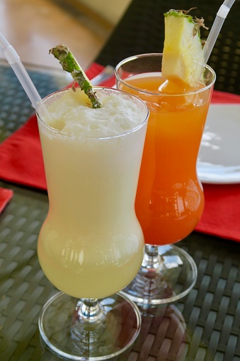 Photo showing alcoholic Piña colada cocktail with  rum, coconut cream and pineapple juice and a non alcoholic Orange Sunset mocktail with grenadine. Both drinks are served in a fiesta hurricane glass at bar, with a clear straw and pineapple garnish.
