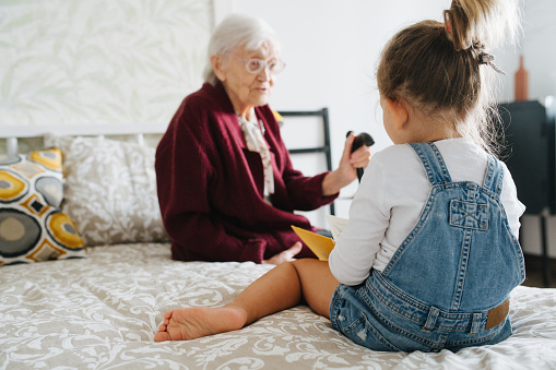 Happy moments with great grandma, senior lady spending quality time with her great granddaughter. Both sitting on a bed, girl is reading book and grandma listens.