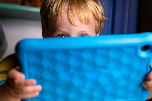 Caucásico niño pequeño juega con su tableta azul mientras se sienta y come en su cocina photo