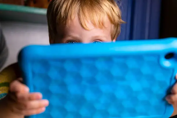 Caucasian Toddler Boy Plays With His Blue Tablet As He Sits And Eats In His Kitchen