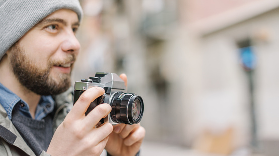 Male casually dressed bearded tourist with a backpack holding retro film photo camera in hands and looking for good angle to shoot. Traveling and freelance concept. Copy space for your text.
