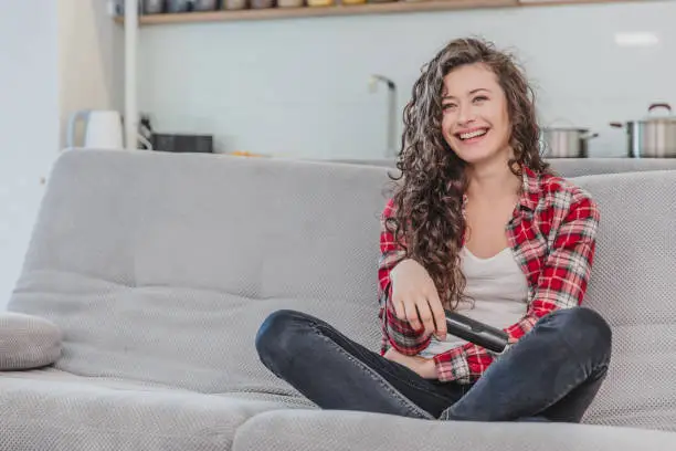 Photo of A beautiful woman watches the TV and is sitting on the couch and holds the remote control in his hand. A brunette in a shirt looks at the TV with long hair.