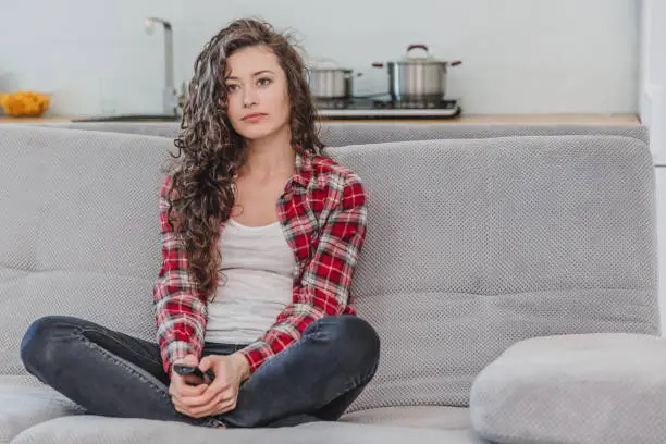 Photo of A beautiful woman watches the TV and is sitting on the couch and holds the remote control in his hand. A brunette in a shirt looks at the TV with long hair.