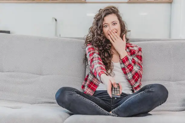 Photo of A beautiful woman watches the TV and is sitting on the couch and holds the remote control in his hand. A brunette in a shirt looks at the TV with long hair.