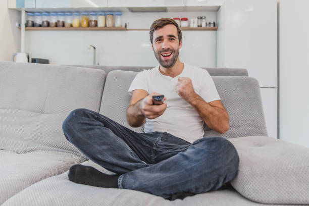istock Beautiful hilarious young man holding a remote control. During this, the TV is watching while sitting on the couch at home. 1140067060
