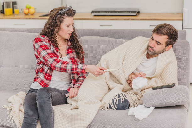 istock A man with a cold sitting on a sofa in a blanket blanket. Checking the temperature. The wife is sitting next to her and helps. Man at home. Sick young man Gray sofa in the room. Unhappy guy. Concept of disease. 1140067062