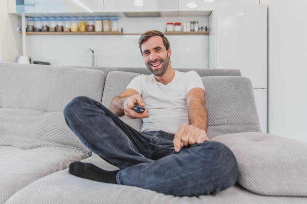 istock Beautiful hilarious young man holding a remote control. During this, the TV is watching while sitting on the couch at home. 1140067199