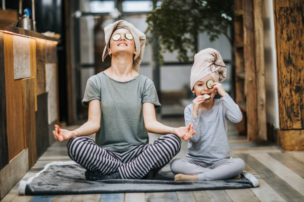 Relaxed mother and daughter exercising Yoga in the morning at home. Mother and little girl taking care of their bodies in the morning while doing Yoga meditation exercises at home. Girl is cheating while eating cucumbers from her eyes and looking at camera. body care and beauty photos stock pictures, royalty-free photos & images