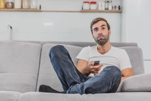 istock Beautiful hilarious young man holding a remote control. During this, the TV is watching while sitting on the couch at home. 1140067268