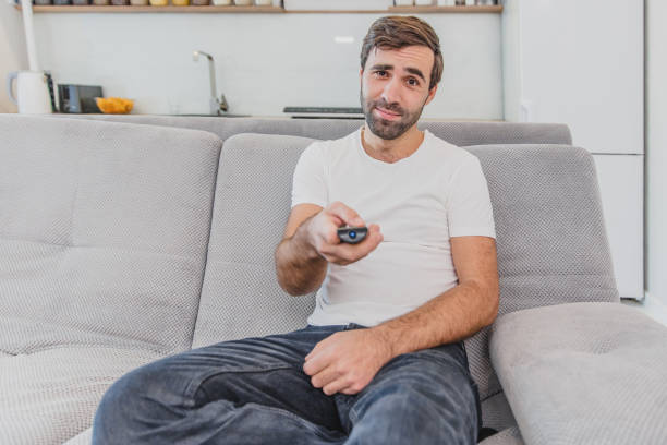 istock Beautiful hilarious young man holding a remote control. During this, the TV is watching while sitting on the couch at home. 1140067273
