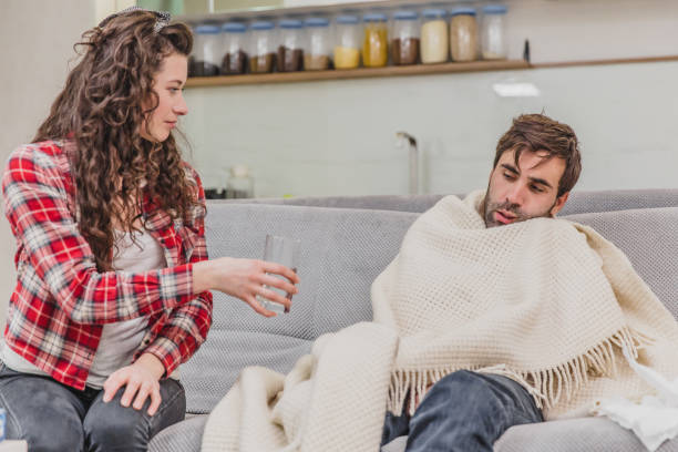 istock A man with a cold sitting on a sofa in a blanket blanket. Checking the temperature. The wife is sitting next to her and helps. Man at home. Sick young man Gray sofa in the room. Unhappy guy. Concept of disease. 1140067276