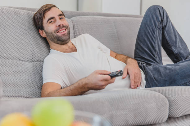 istock Beautiful hilarious young man holding a remote control. During this time he looks at the camera, smiles while sitting on the couch at home. 1140067320