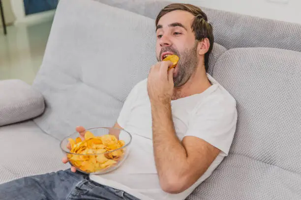 Photo of The guy in the T-shirt sits on the couch, eats chips and watches the TV. While holding the remote in his hands. Personality at home.