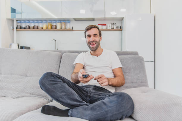 istock Beautiful hilarious young man holding a remote control. During this, the TV is watching while sitting on the couch at home. 1140067374