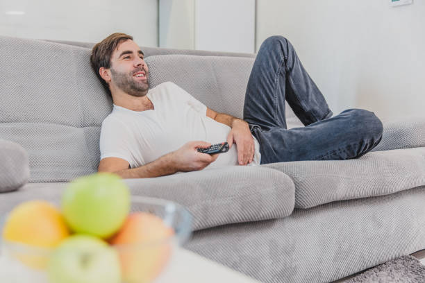 istock Beautiful hilarious young man holding a remote control. During this, the TV is watching while sitting on the couch at home. 1140067397