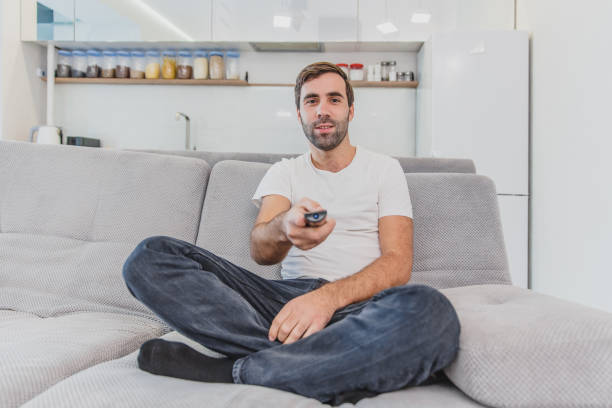 istock Beautiful hilarious young man holding a remote control. During this, the TV is watching while sitting on the couch at home. 1140067421