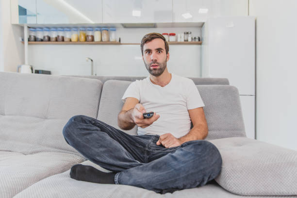 istock Beautiful hilarious young man holding a remote control. During this, the TV is watching while sitting on the couch at home. 1140067472