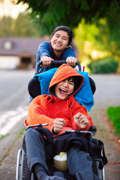 Sister pushing disabled little brother in wheelchair around neighborhood stock photo