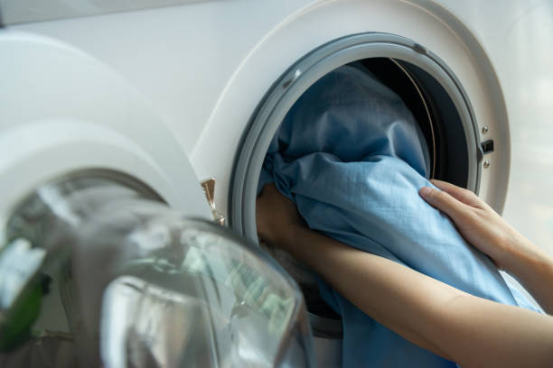 Open door in washing machine with blue bed sheets inside close up Woman’s hand loading dirty blue bed sheets in a white washing machine. bedding stock pictures, royalty-free photos & images