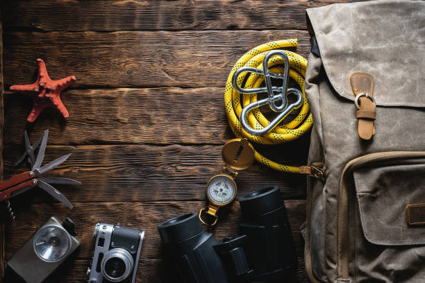 Travel or adventure flat lay background with copy space. A backpack, photo camera, flashlight, multitool knife, rope, carbines, binoculars, compass and a starfish on a brown wooden table.