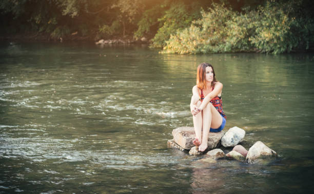 A caucasian girl in summer shorts sitting on a stone in the river