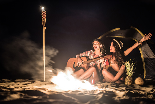 Two happy female friends camping on a beach next to the fire.