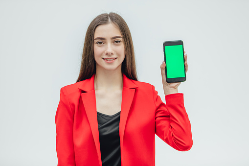 Young pretty pretty girl wearing a red jacket. Stays on a gray background. During this time, holding a black phone with one hand in your hand. Green screen