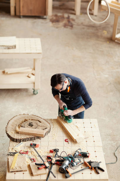 istock Professional carpenter sanding wooden plank with flatbed sander machine in his loft studio, his workbench covered with tools, wood shavings and sawdust 1140094725
