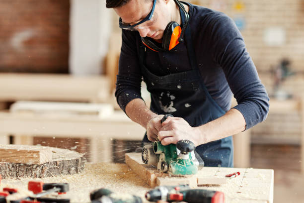 istock Young woodworker in protective eyeglasses sanding wooden plank with electric sanding machine, sawdust flying away 1140094727