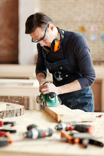 istock Young woodworker sanding wooden plank with flatbed sander machine, his workbench covered with tools, wood shavings and sawdust 1140094729
