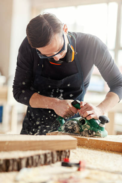 istock Young woodworker sanding wooden plank with electric flatbed sander, sawdust flying away 1140094749
