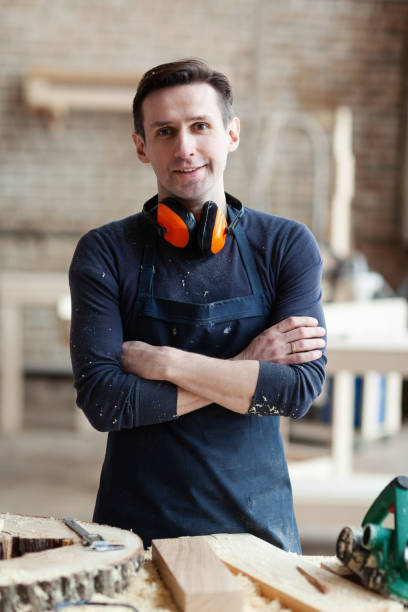 istock Young smiling woodworker in apron with crossed arms looking at camera while standing at workbench with pieces of wood and electric plane on it 1140094756