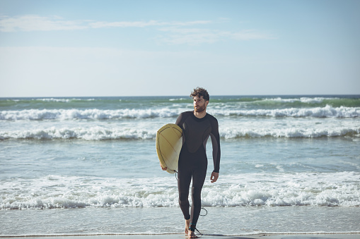 Front view of young Caucasian male surfer with a surfboard walking on a beach on sunny day. He is holding his surfboard